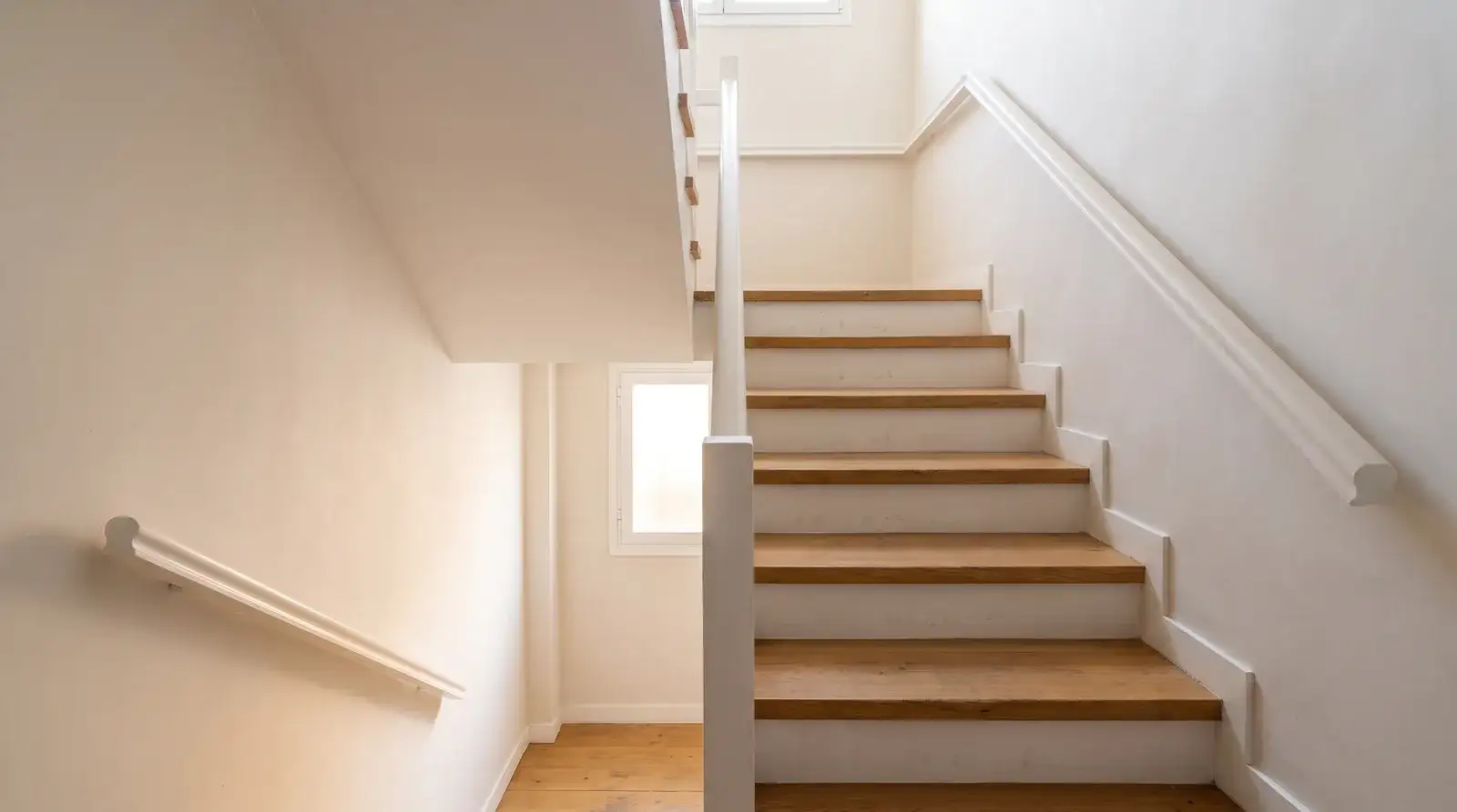 Escalera interior de una vivienda con peldaños de madera de roble, contrahuellas blancas y pasamanos renovado bajo luz natural